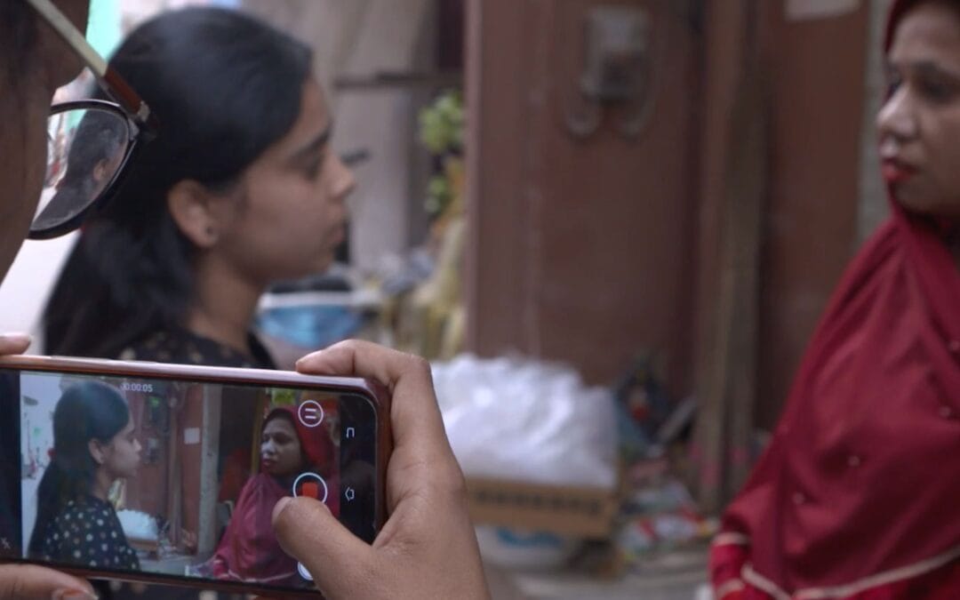 An Indian woman uses a mobile phone to film a younger Indian woman interviewing another Indian woman outside on a street